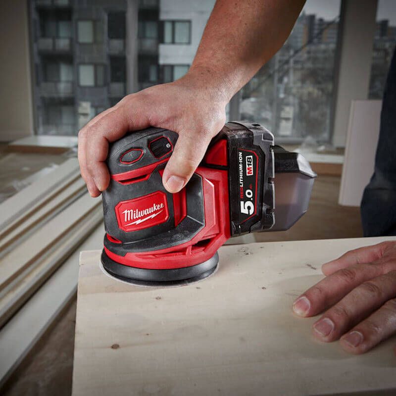 A person using the MILWAUKEE® M18 125mm Random Orbital Sander to smooth a wooden surface, showcasing its dust extraction feature and cordless operation in a professional workspace. A person using the MILWAUKEE® M18 125mm Random Orbital Sander to smooth a wooden surface, showcasing its dust extraction feature and cordless operation in a professional workspace.